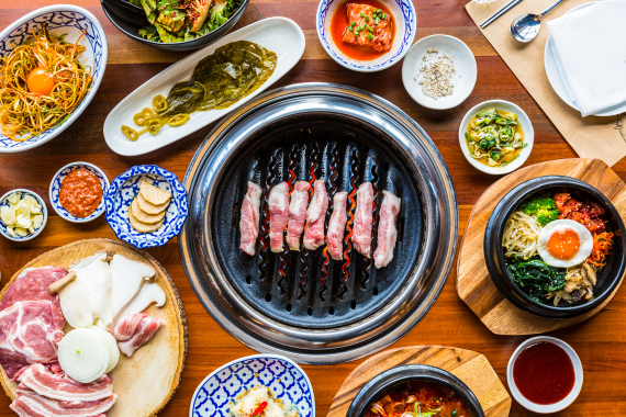Gevulde tafel met een grill in het midden waarop dungesneden vlees gegrild wordt. Errond staan kommetjes met Koreaanse bijgerechten zoals kimchi, lente-uisalade, bibimbap, een stoofpotje en rauw vlees en champignons voor op de grill.
