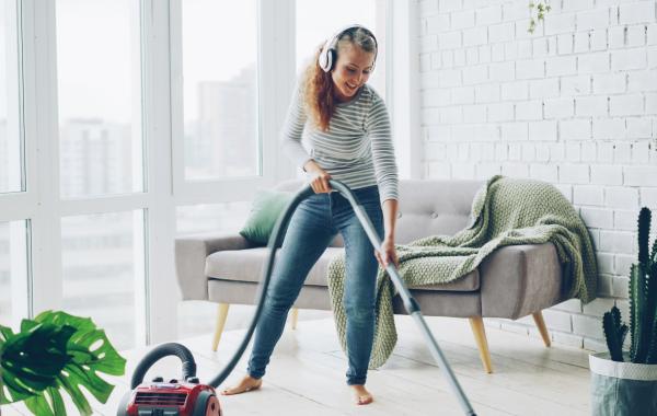 Vrouw met koptelefoon in lichtrijke, witte woonkamer met planten, die lachend huis schoonmaakt met stofzuiger